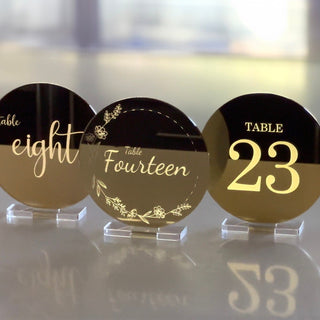 Three backlit gold mirror acrylic table numbers. The personalised design is engraved, leaving the acrylic translucent. The foreground lighting is dim, and the background is bright, illuminating the engraved design.