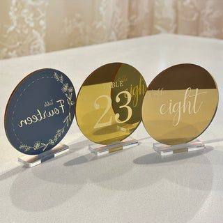 Three circular table numbers made from gold mirror acrylic. One of these is facing away from the camera showing the slate grey backing colour of the mirror material. The design is etched into the grey opaque backing, making the design glow.